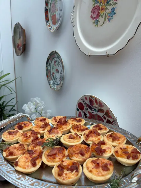 Bandeja prateada com mini quiches de queijo e bacon, ramos de tomilho e pratos decorativos na parede ao fundo
