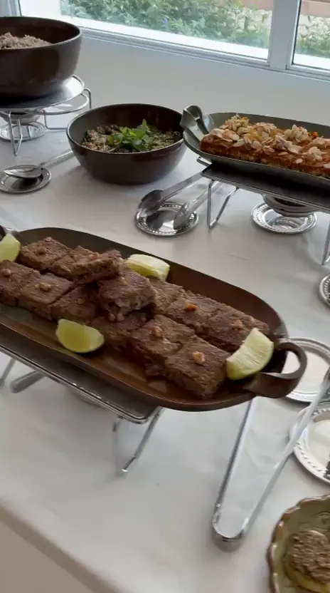 Buffet de dia com kibe assado com limão, charutinho e tigelas de pastas sobre toalha branca